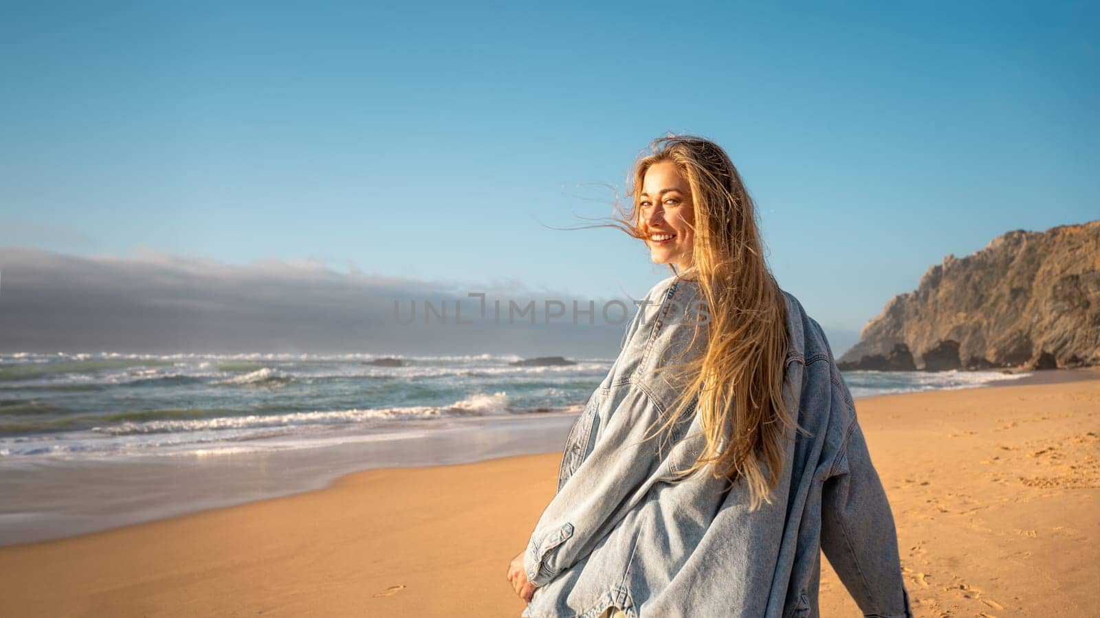 Woman walking ocean beach and turns around, smiling at the camera. Happy female traveler dressed denim shirt walk sandy sea beach, holiday vacation at summer.