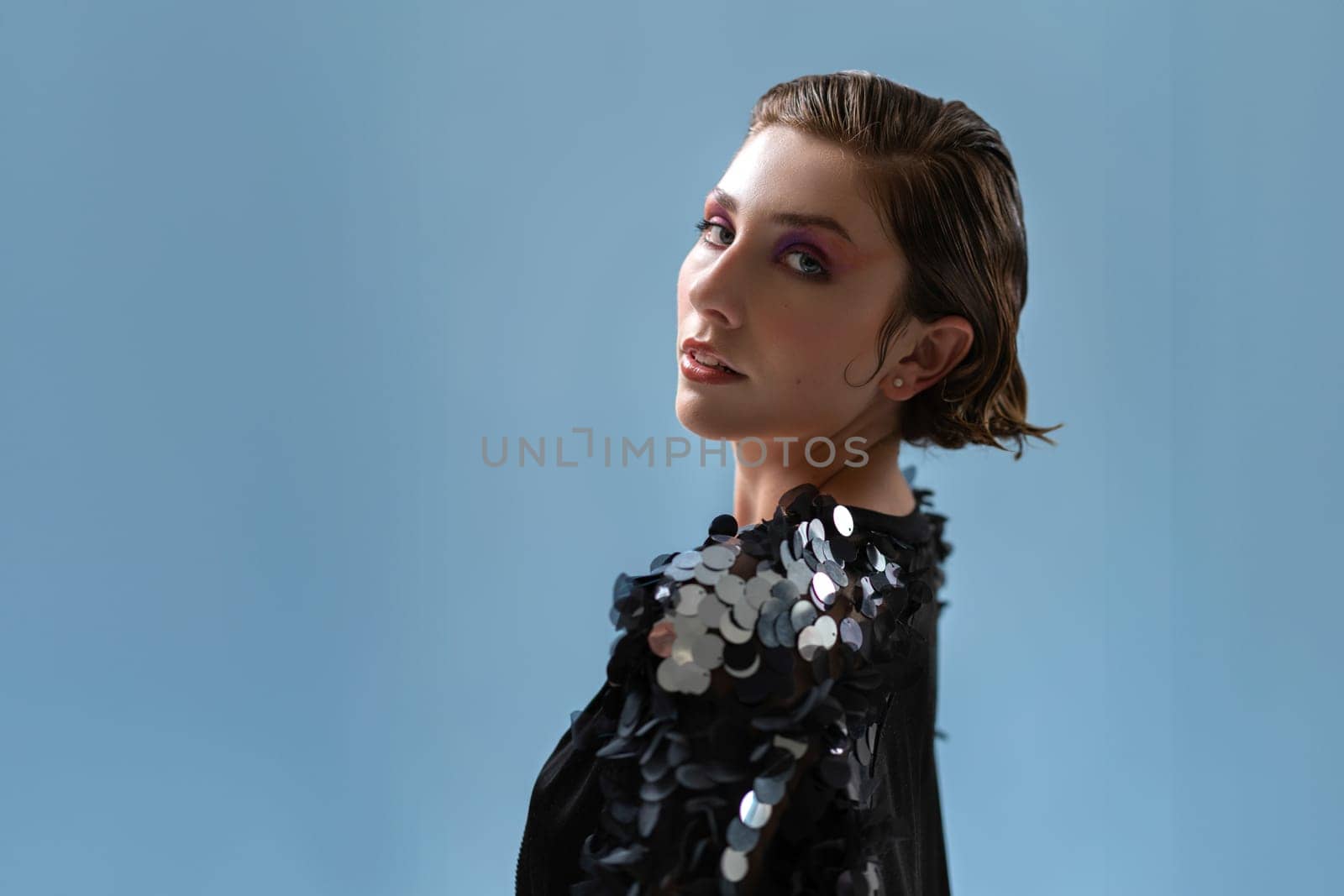 Fashionable confident woman wearing stylish black jacket with paquettes, standing isolated on blue studio background looking at camera over shoulder. Studio fashion portrait