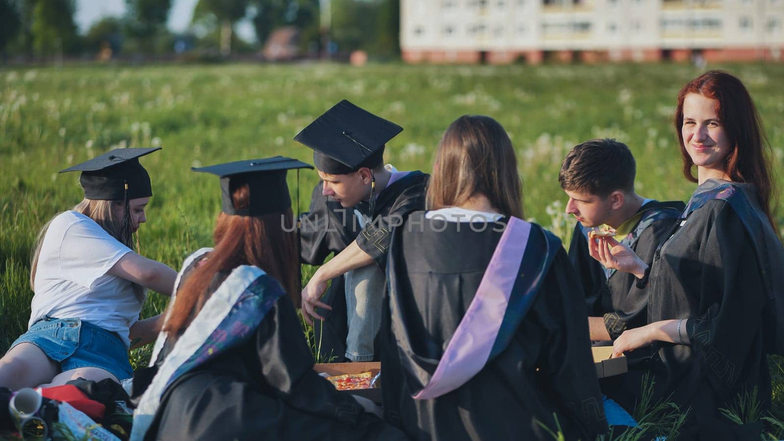 Graduates in black suits eating pizza in a city meadow