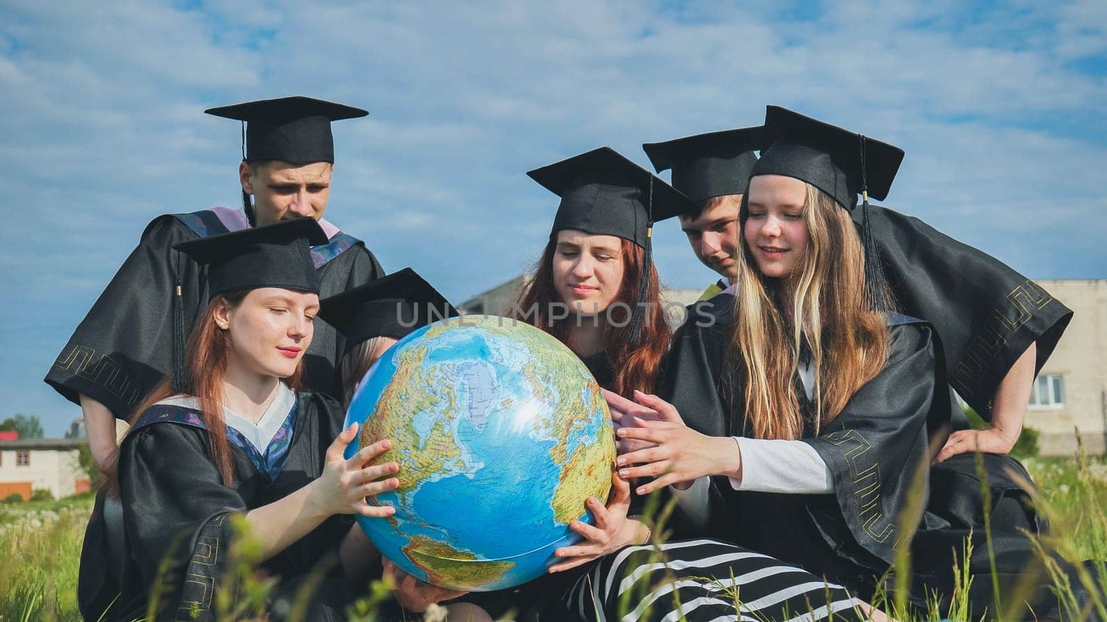 Graduates in black robes examine a geographical globe sitting on the grass