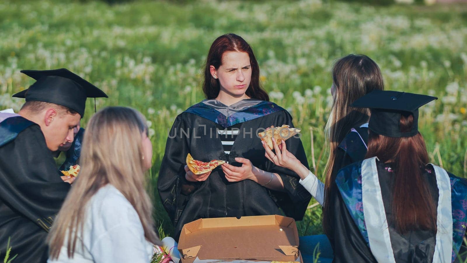 Graduates in black suits eating pizza in a city meadow. by DovidPro