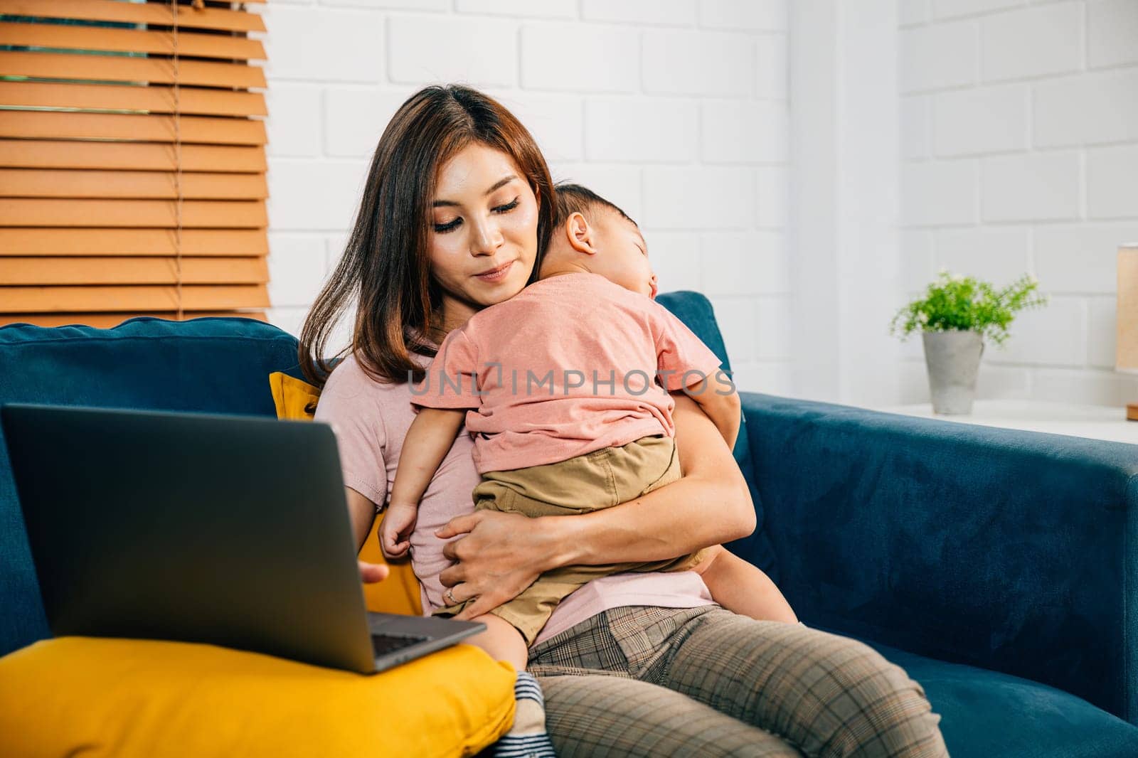 Balancing motherhood and work a businesswoman types on her laptop as her baby daughter peacefully naps on the sofa. This portrait captures their loving bond.
