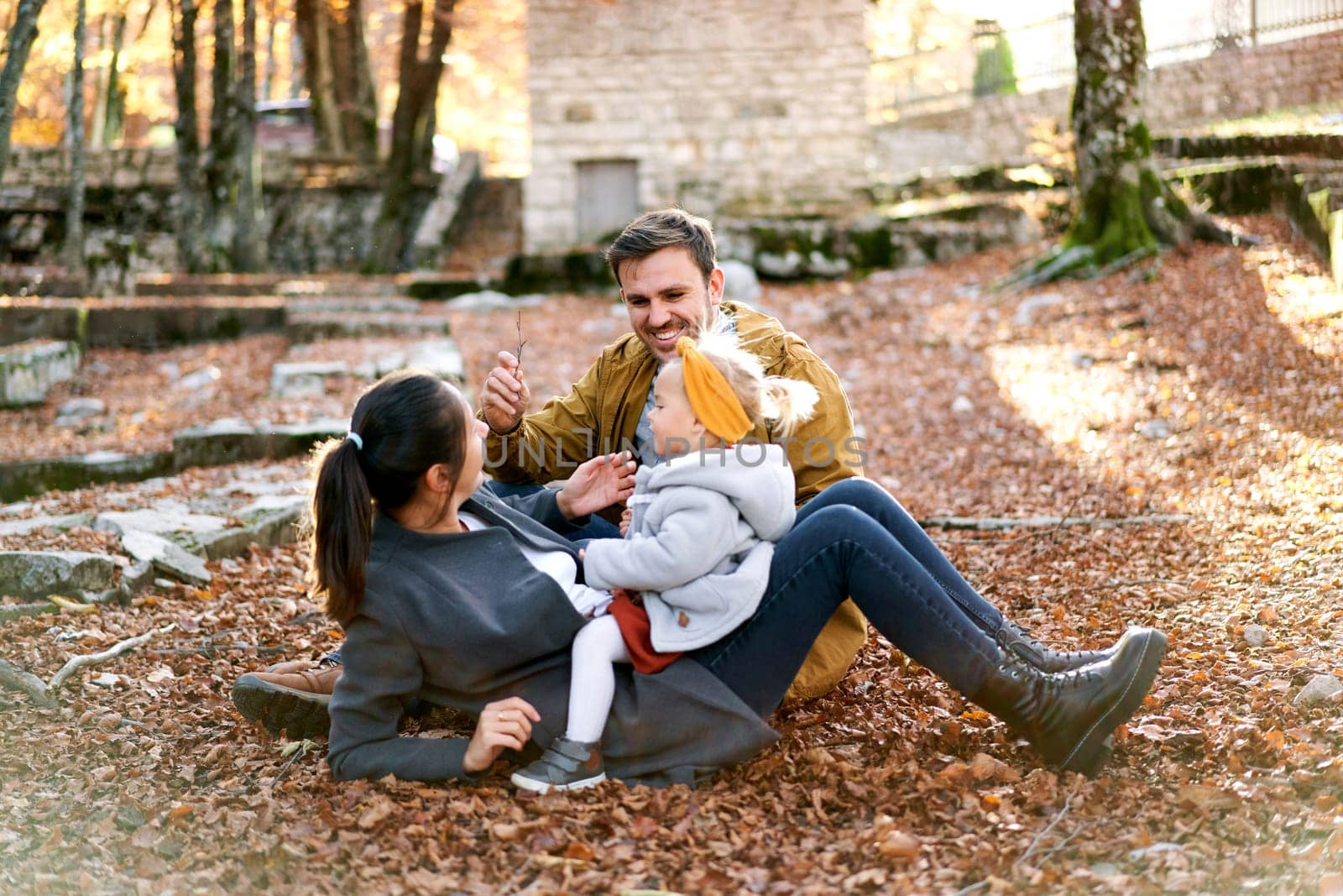 Smiling dad sits near mom with a little girl on her belly on fallen leaves in the forest. High quality photo