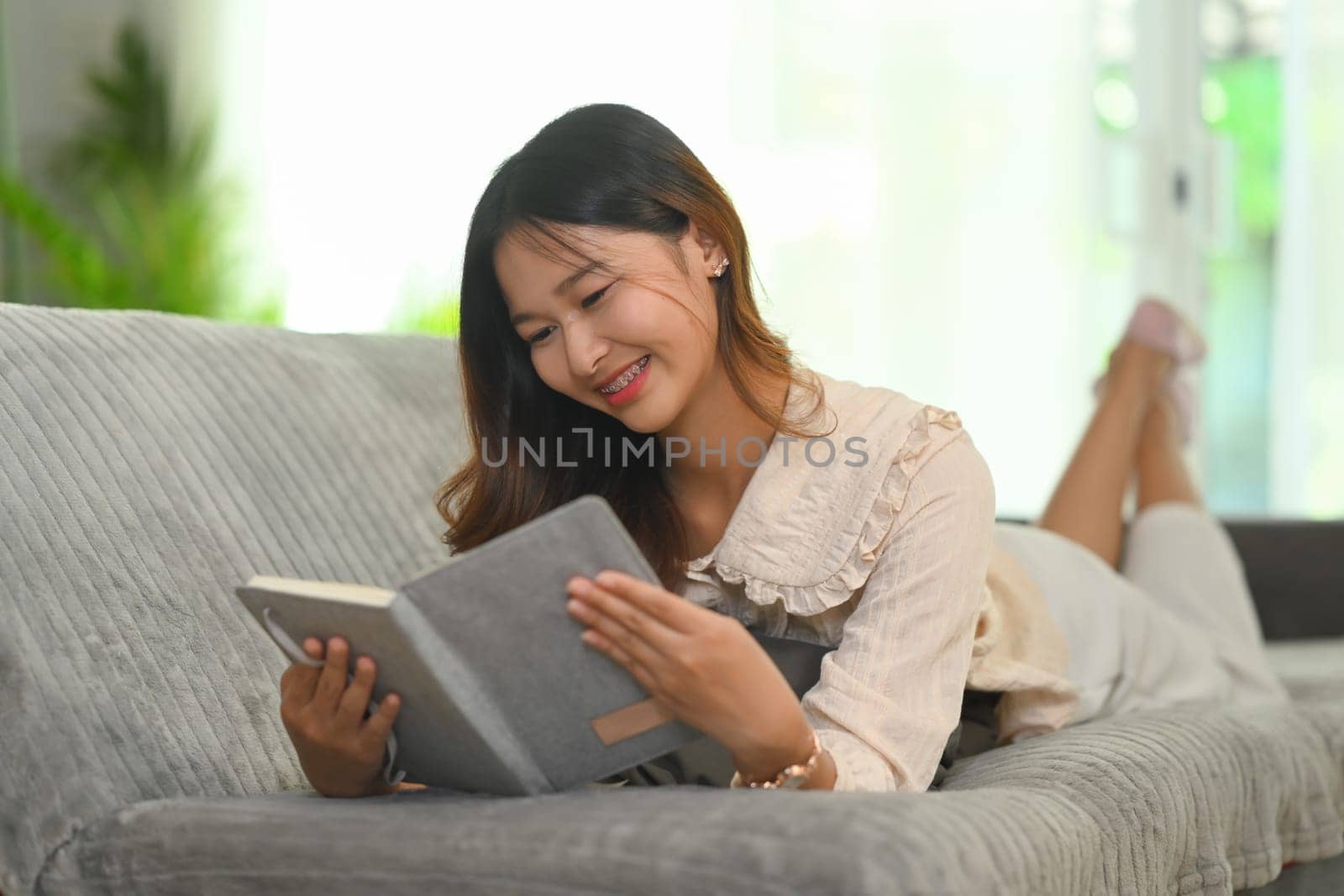 Happy young woman lying on cozy sofa and reading book. People, leisure and lifestyle concept.