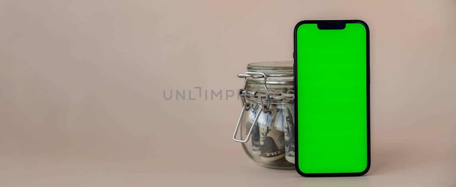 Banner Vertical Green screen on modern mobile phone in background of glass jar full of American currency dollar banknotes on beige background. Cope space for text. Advertisement for application website. Concept of money economy banks and finances