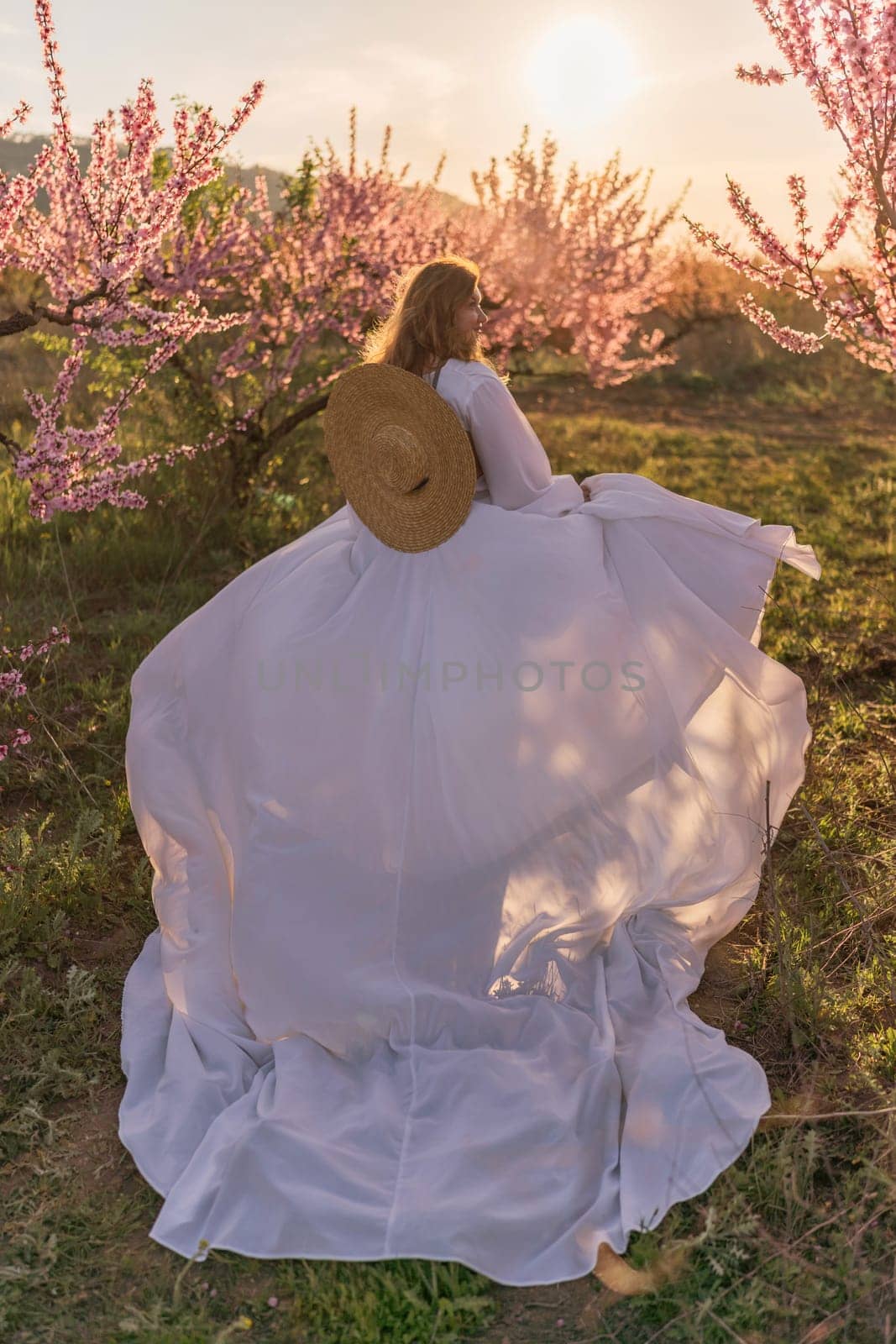 Woman blooming peach orchard. Against the backdrop of a picturesque peach orchard, a woman in a long white dress and hat enjoys a peaceful walk in the park, surrounded by the beauty of nature