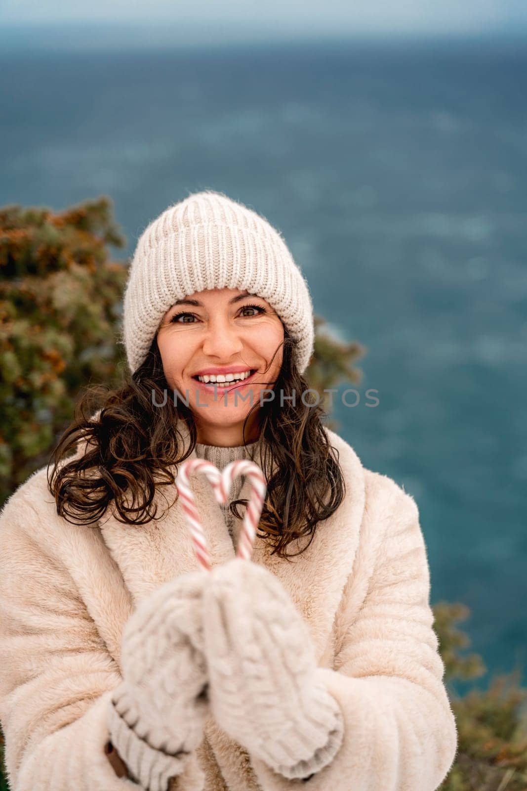 Woman candy sea. Smiling woman in knitted hat, mittens and beige coat holding lollipops candy canes in her hands in shape of heart against the backdrop of the sea. by Matiunina