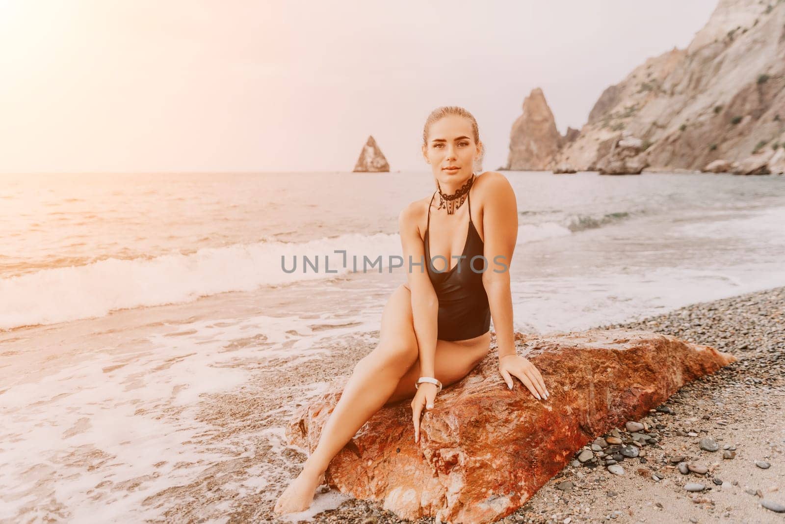 Woman travel portrait. Happy woman with long hair looking at camera and smiling. Close up portrait cute woman in black bikini posing on a red volcanic rock on the sea beach by panophotograph