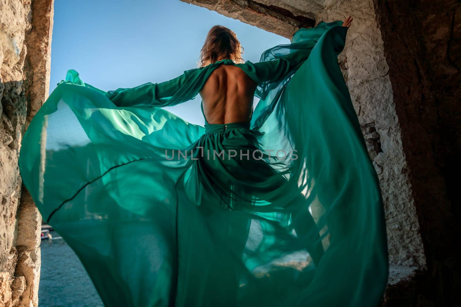 Rear view of a happy blonde woman in a long mint dress posing against the backdrop of the sea in an old building with columns. Girl in nature against the blue sky