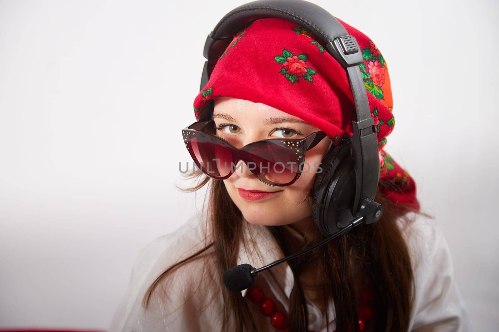 Portrait of young girl in red scarf, large headphones with microphone and black glasses. Woman who is radio or television presenter in the workplace. Funny female telecom operator. Freelancer at work by keleny