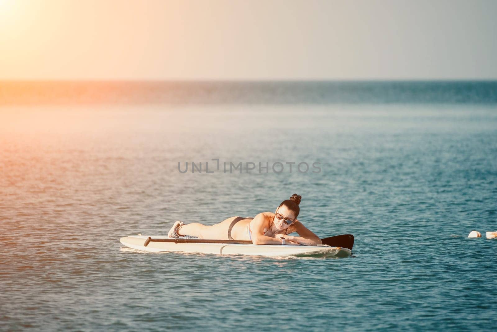 Sea woman sup. Silhouette of happy middle aged tanned woman in rainbow bikini, surfing on SUP board, confident paddling through water surface. Idyllic sunset. Active lifestyle at sea or river. by panophotograph