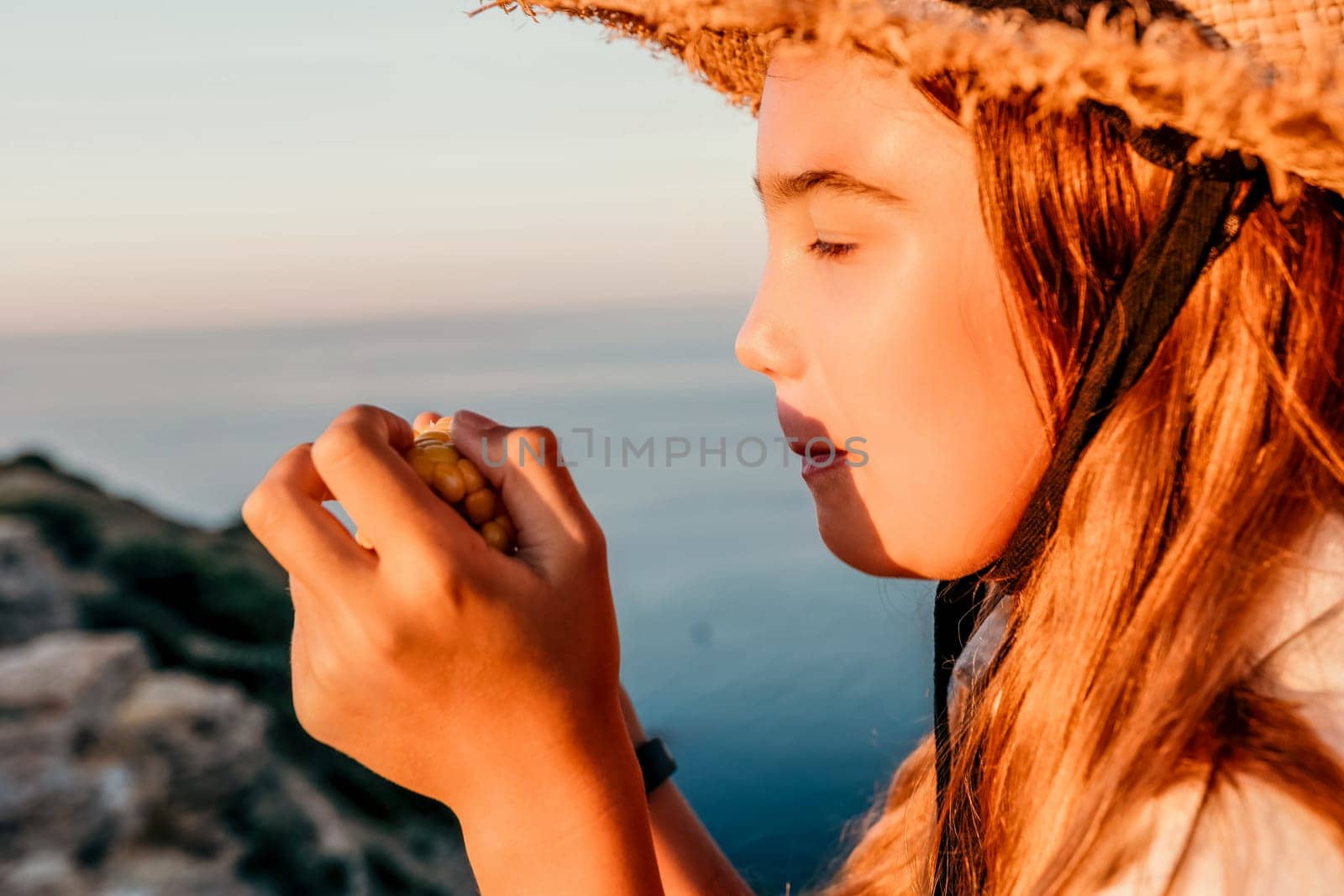 Happy girl eating corn. Summer snacking on the sea. Portrait of young beautiful girl in straw hat eating grilled corn while sitting by the sea on sunset time. Close up. Selective focus by panophotograph