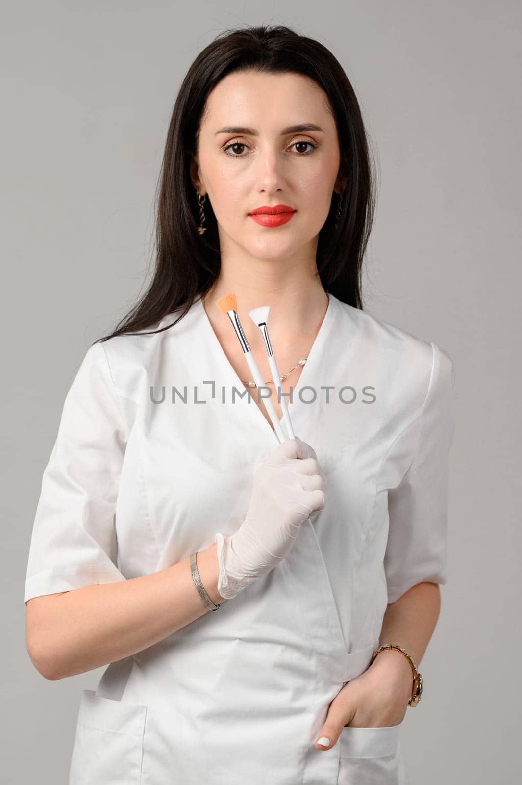 the girl cosmetologist holds two brushes in her hands on a white background, the girl is a beautician in white disposable gloves.