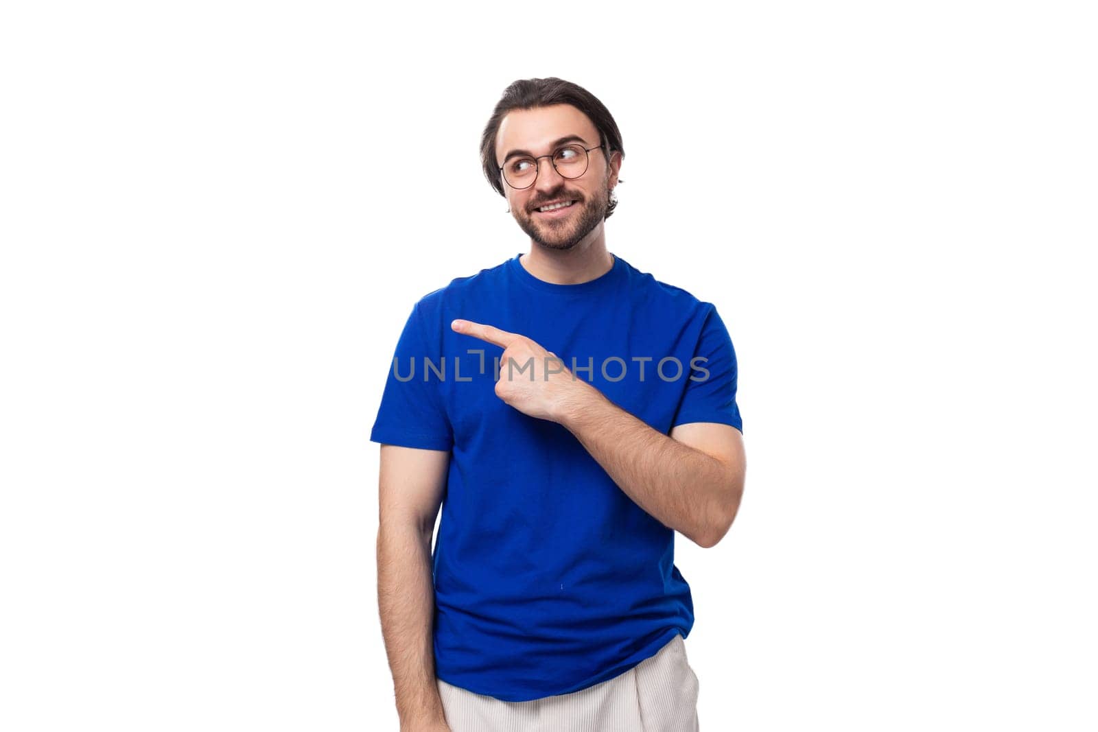 young smart brunette man with a beard dressed in a blue t-shirt points with his hand to the space for advertising on a white background by TRMK