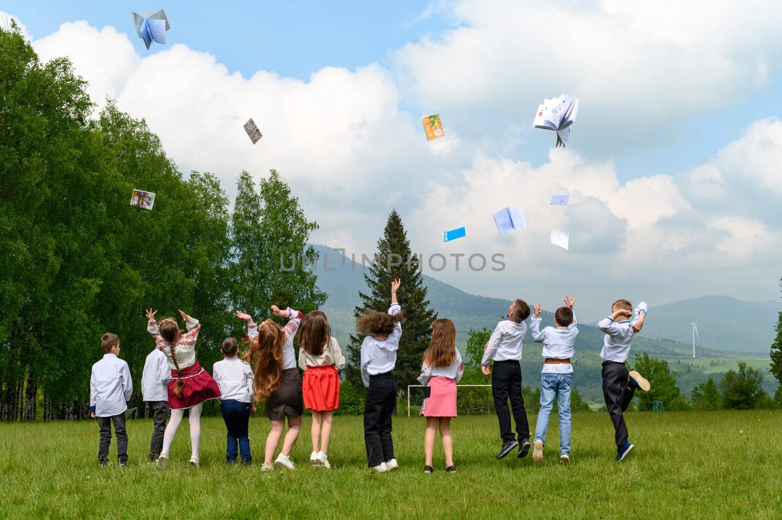 a group of young schoolchildren throw their notebooks up, mountains and forest in the background.