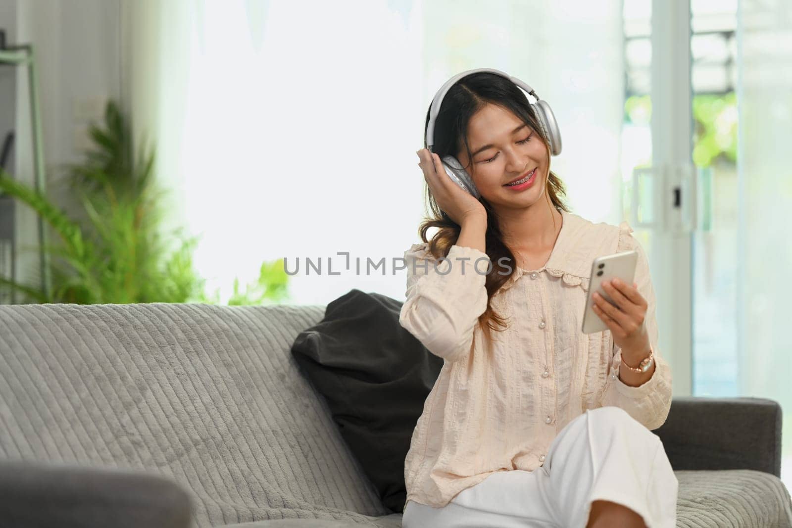 Cheerful young woman listening to music in headphones and using smartphone on couch