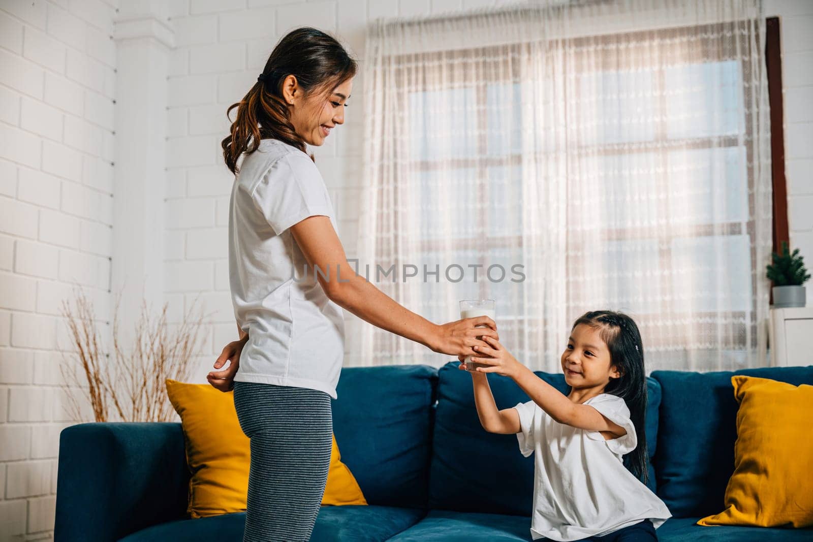 In a cozy living room an Asian mother shares a glass of milk with her daughter on the sofa celebrating the joy of togetherness promoting the importance of calcium and nurturing their strong bond.