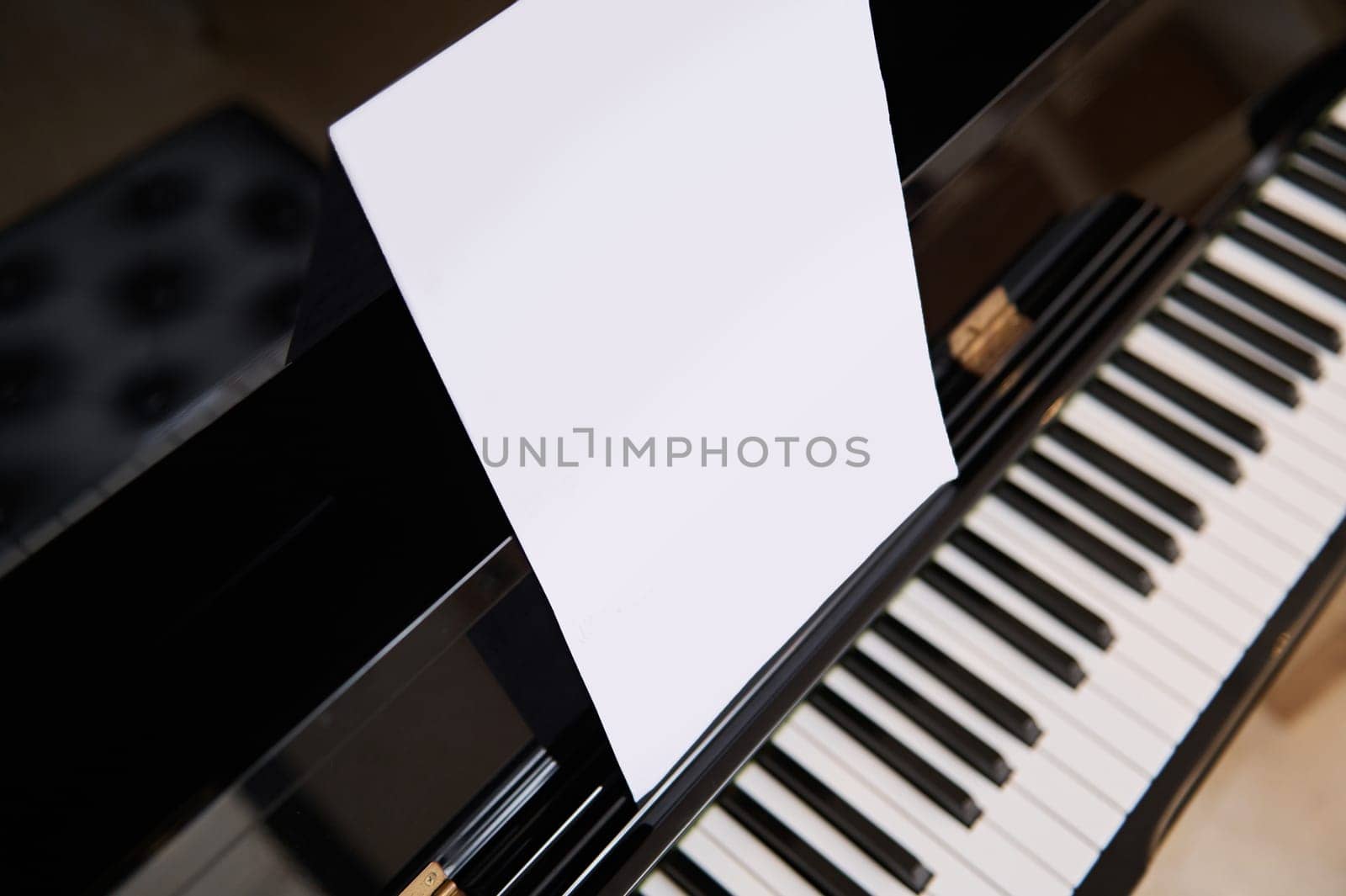 Close-up of old black wooden piano with ivory and ebony piano keys and white paper sheet with copy space for notes or advertising text. View from above