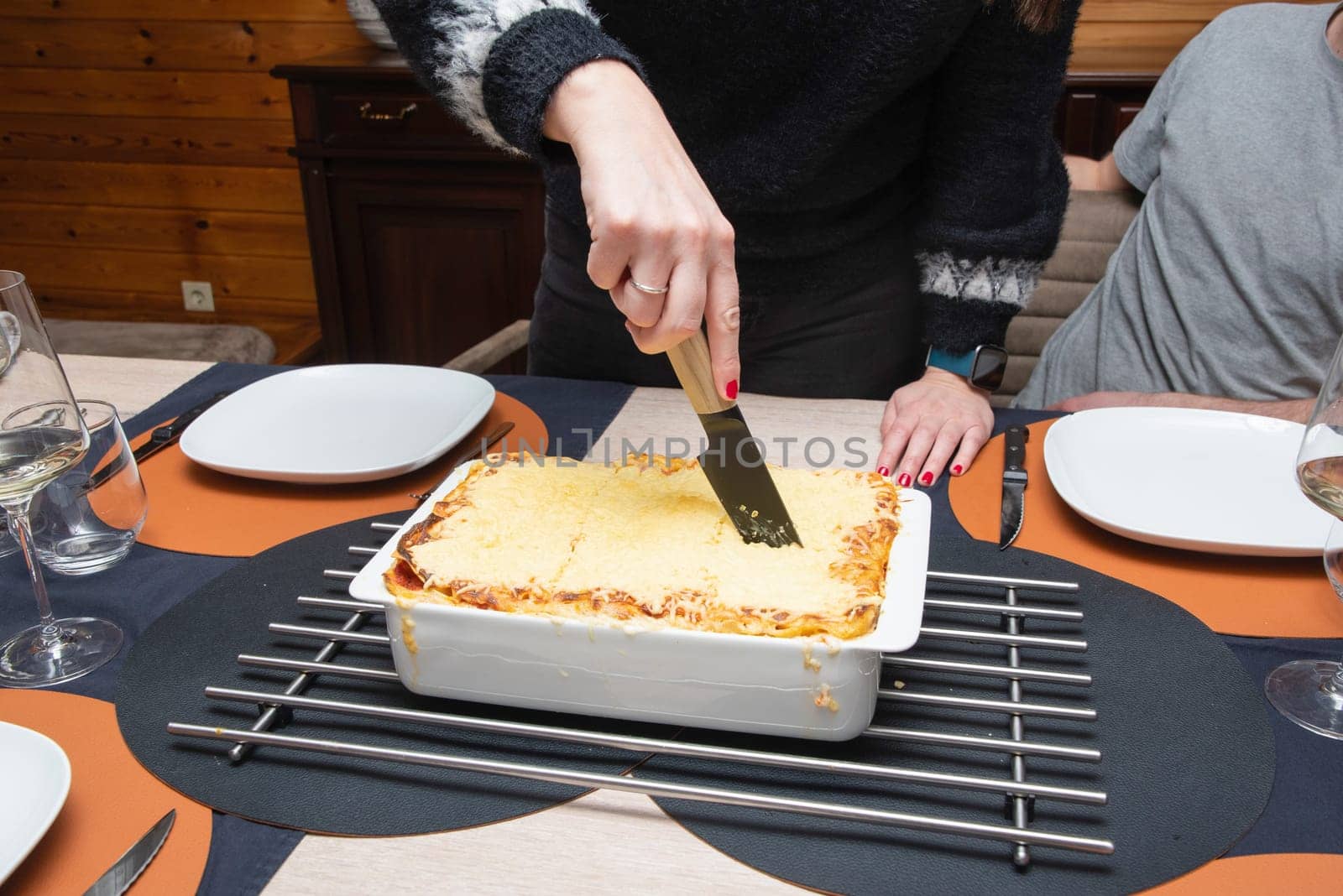 Woman cut with knife hot tasty home baking lasagna in ceramic casserole dish, whole family sit at the table, High quality photo