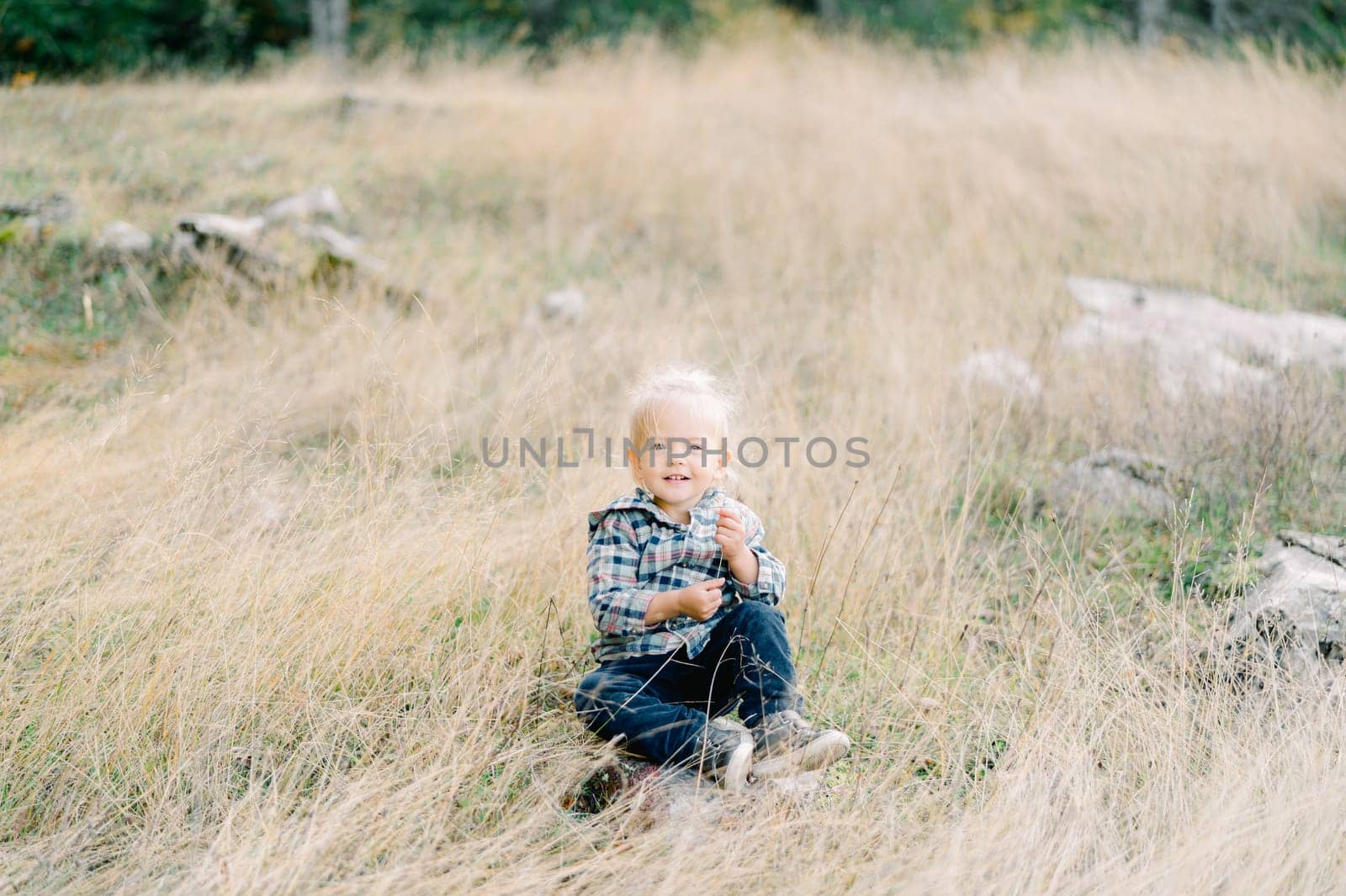 Little smiling girl sits in dry grass on a stone and plays with a blade of grass. High quality photo