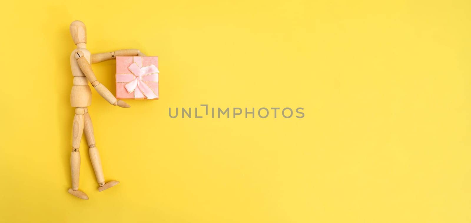 Wooden figurine holding a pink gift box. Yellow background, copy space by darksoul72