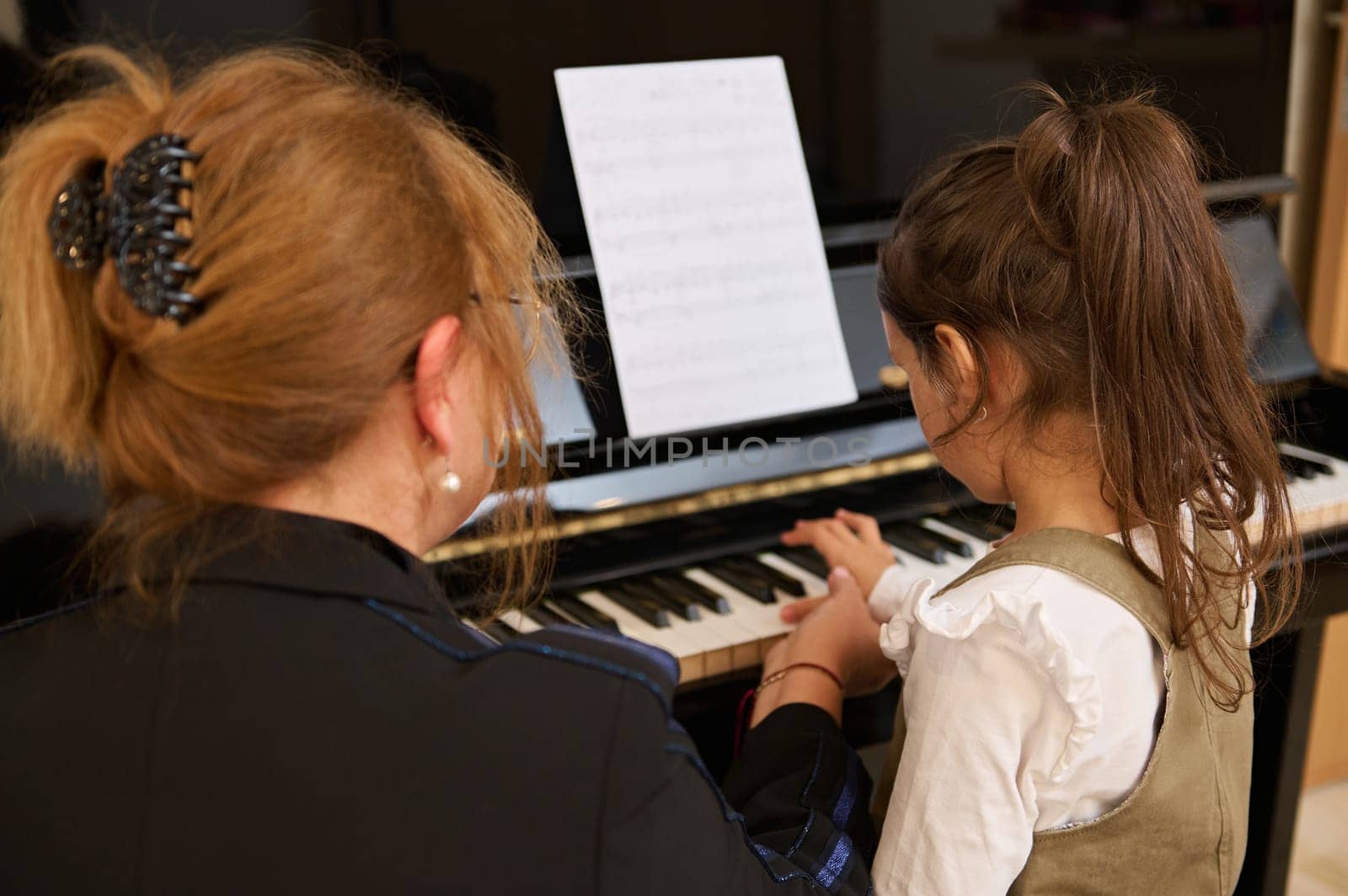 Rear view of a redhead mature female teacher, a professional pianist musician and little child girl student playing together classic melody on old grand piano during individual music lesson at home