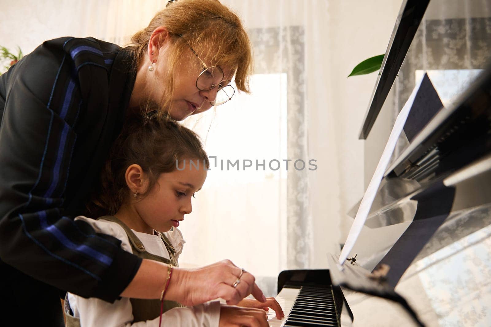 Closeup side view of child touching piano keys under the guidance of a musician pianist teacher explaining piano lesson by artgf