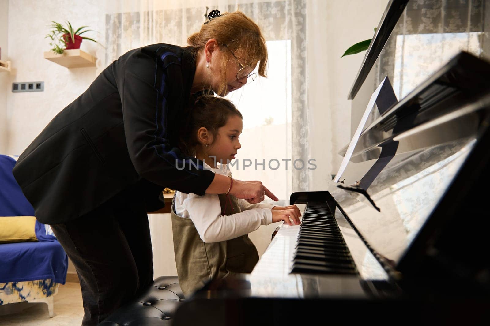 Mature woman pianist teacher giving piano lesson to a little school girl, teaching the position of fingers on piano keys by artgf
