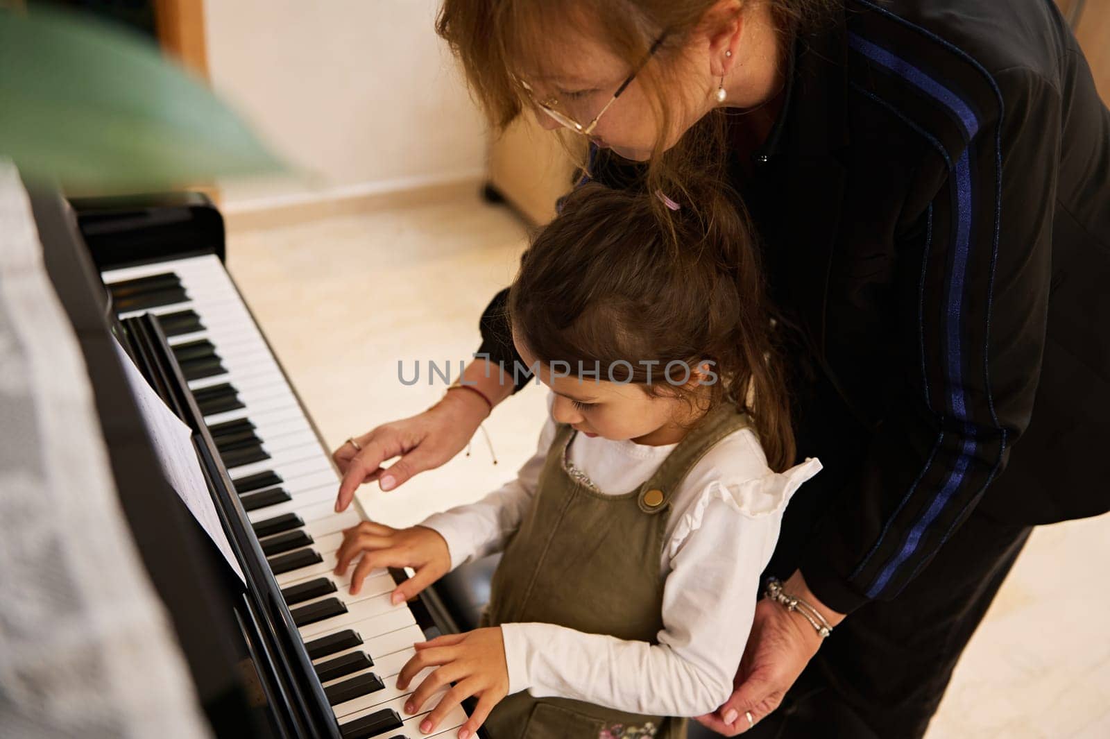 View from above of a female musician teacher and little kid girl touching the piano keys, explaining the true positions of fingers while learning play piano and capturing the rhythm of music