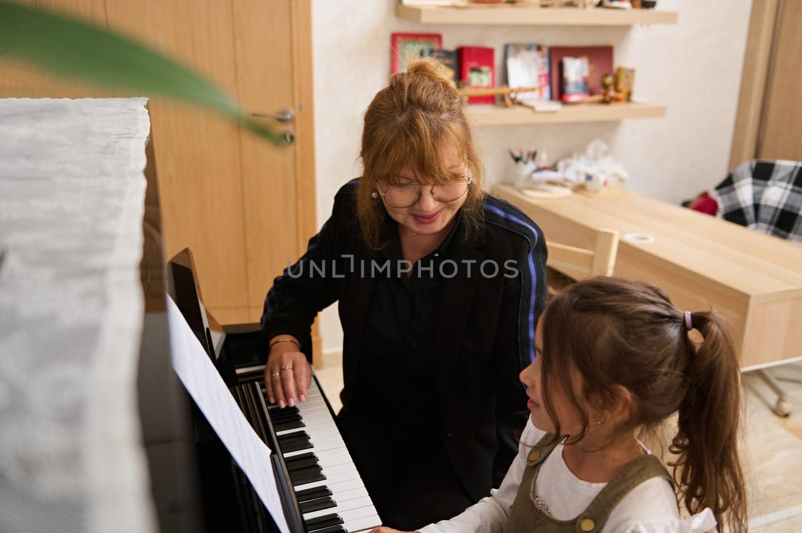 Beautiful mature woman, pianist teacher giving music lesson to a smart child girl, teaching her playing on grand piano by artgf