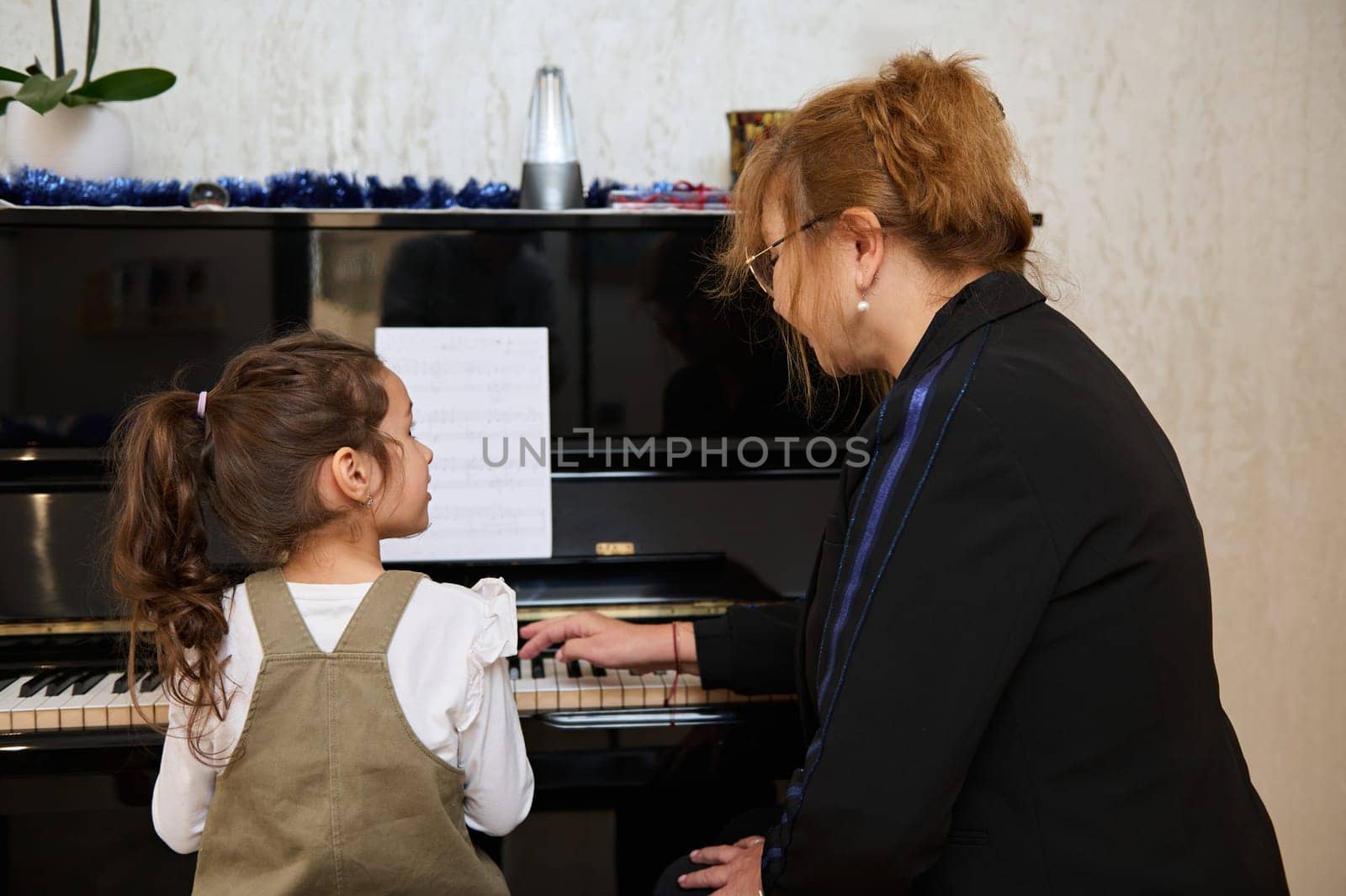 Rear view mature woman, pianist teacher giving music lesson to a smart child girl, teaching her playing on grand piano by artgf
