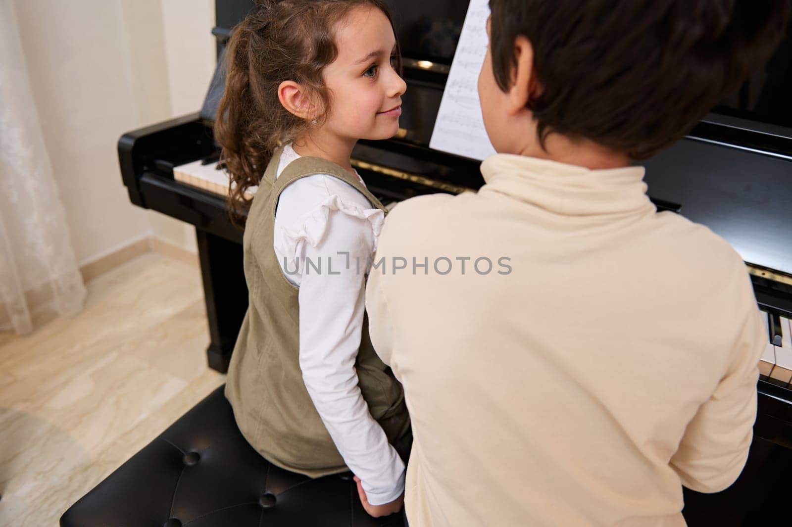 Rear view of two kids playing grand piano together, creating rhythm of classical melody during individual music lesson indoor.