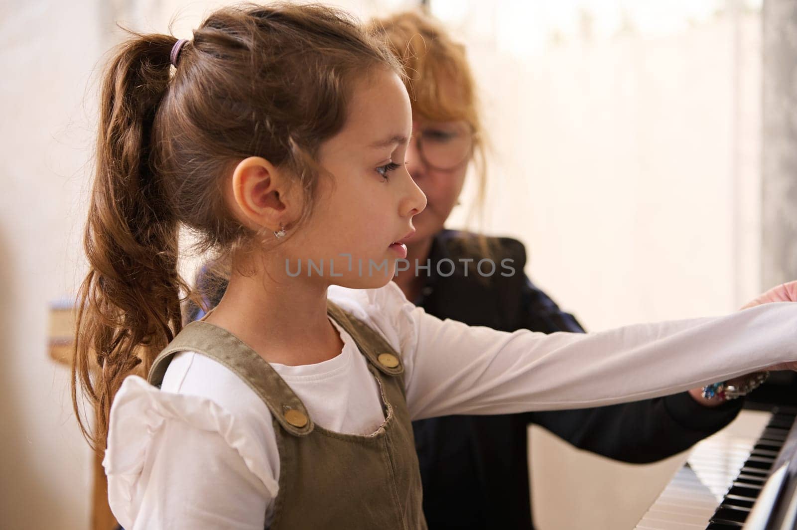 Close-up side portrait of a Caucasian beautiful little child girl, focused engaged on learning playing grand piano with female teacher musician and pianist during individual music lesson at home