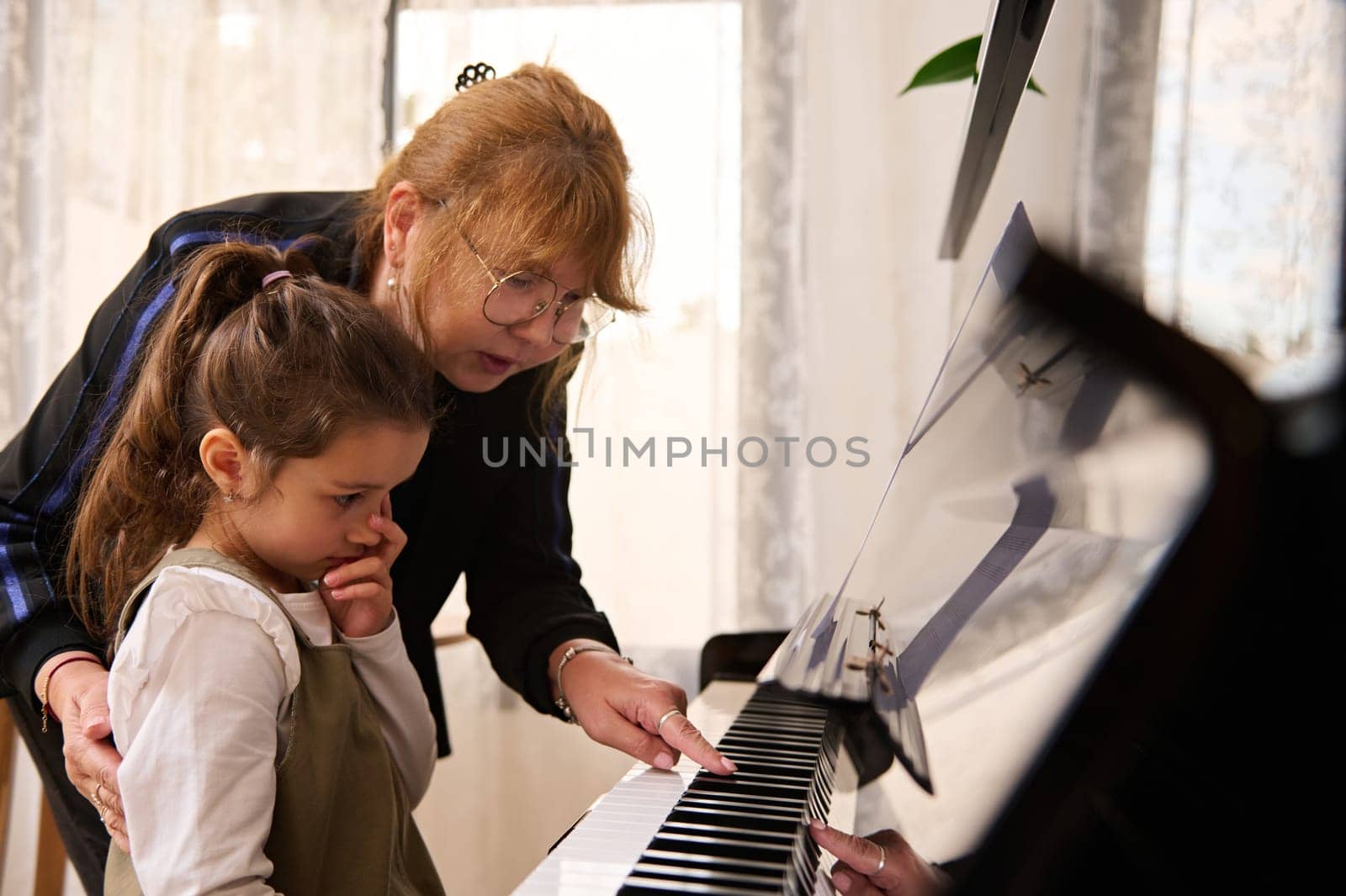 Authentic portrait of a little girl having piano lesson with female teacher musician at home, learning music during individual lesson, touching piano keys, performing classic melody at home interior