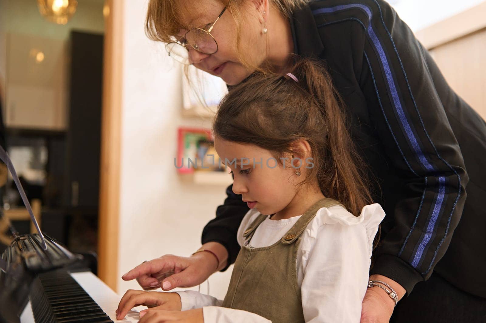 Closeup Caucasian elementary age child, a charming little student girl sitting at piano while a female teacher pianist teacher her, giving music lesson at home. Kids development and entertainment