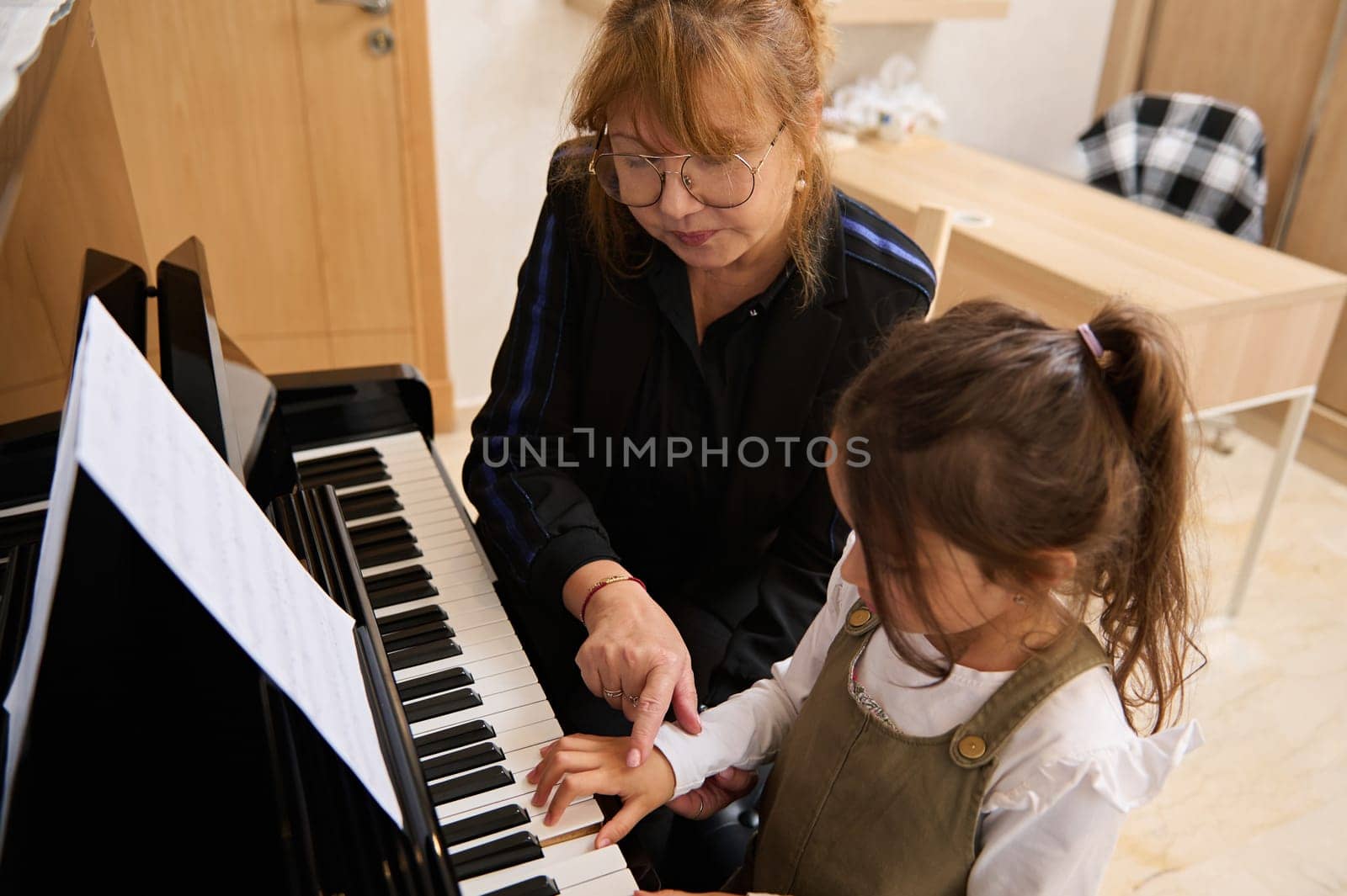 Charming schoolgirl in casual clothes, learning music at home, playing grand piano under the guidance of a music teacher at home during individual lesson