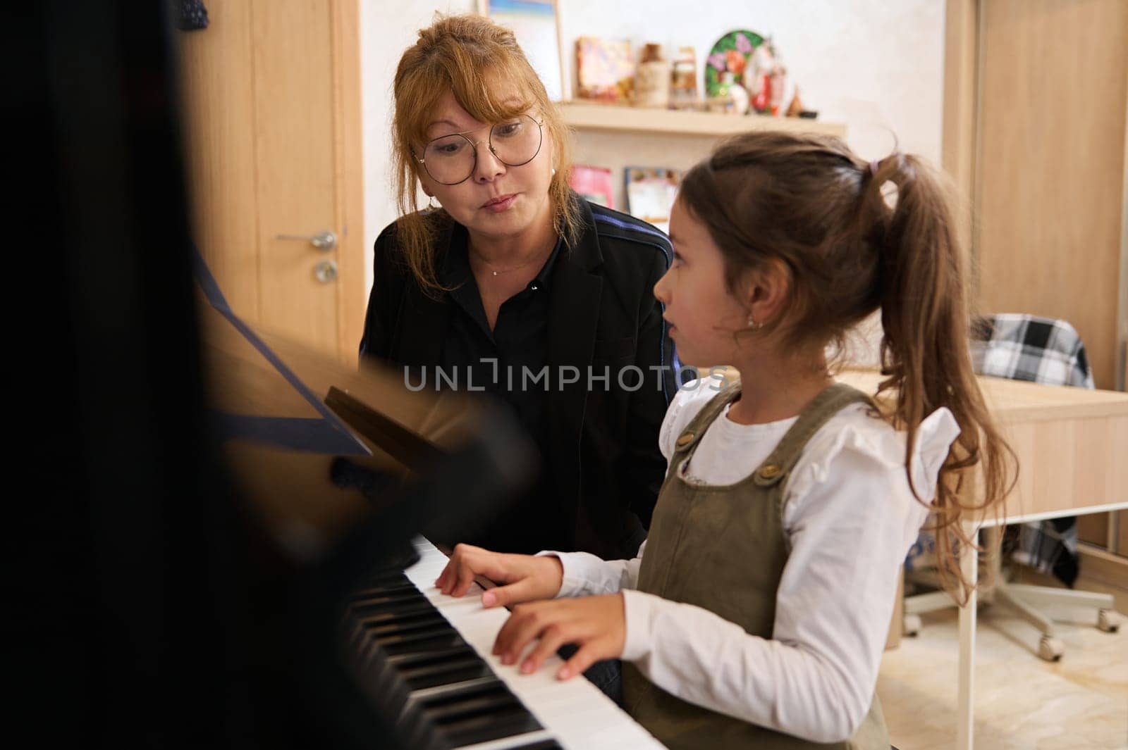 Beautiful little child girl sitting at piano near her teacher pianist musician, learning music during individual music lesson at home