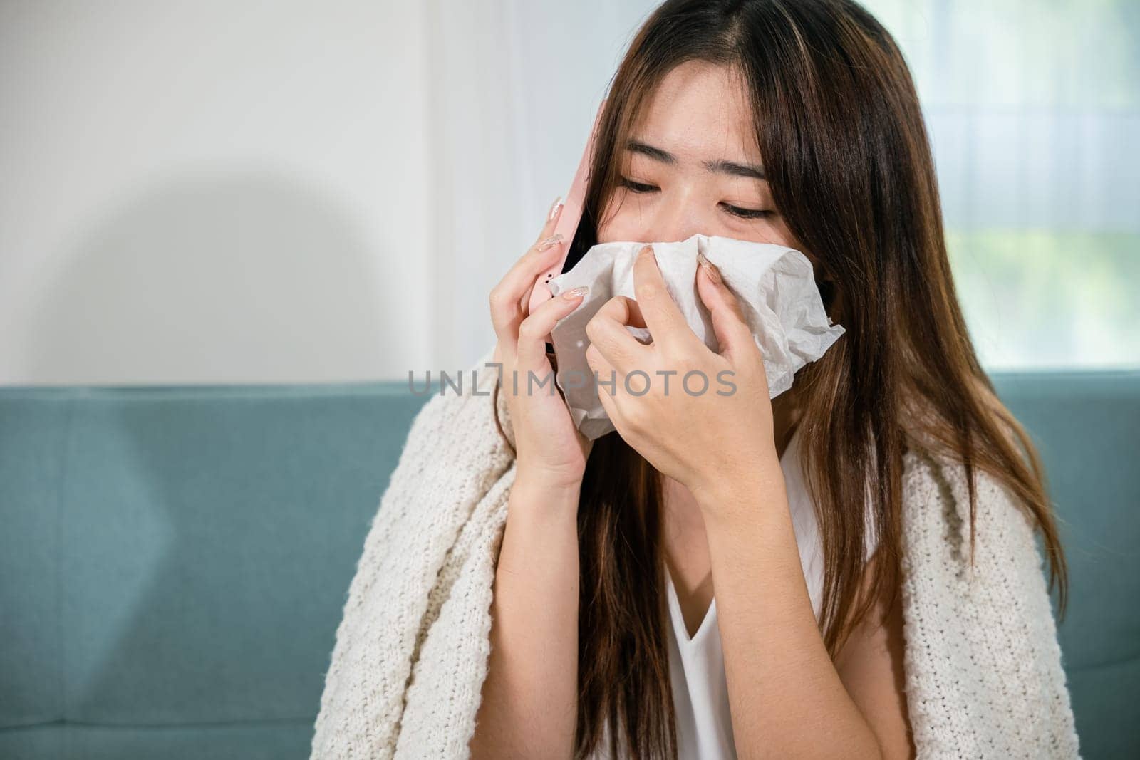 Asian young woman sick sitting down on sofa with cold blowing her nose hold tissues and talking on cellphone to doctor employer while asking about sick leave with mobile phone in living room at home
