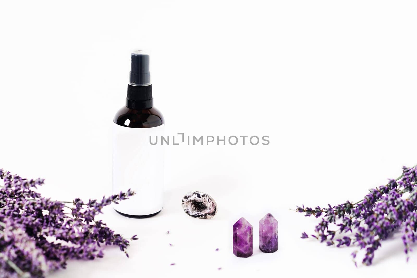 Glass spray bottle on white background surrounded by lavender bouquets and the crystal of quartz. Energy of stones for your home.