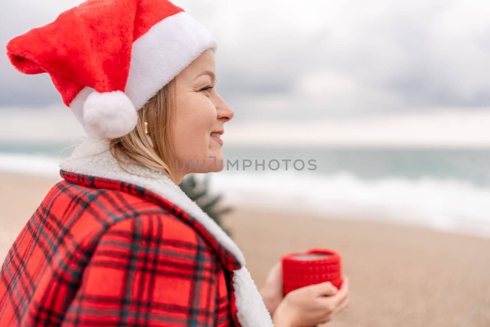 Sea Lady in Santa hat plaid shirt with a red mug in her hands enjoys beach with Christmas tree. Coastal area. Christmas, New Year holidays concep.