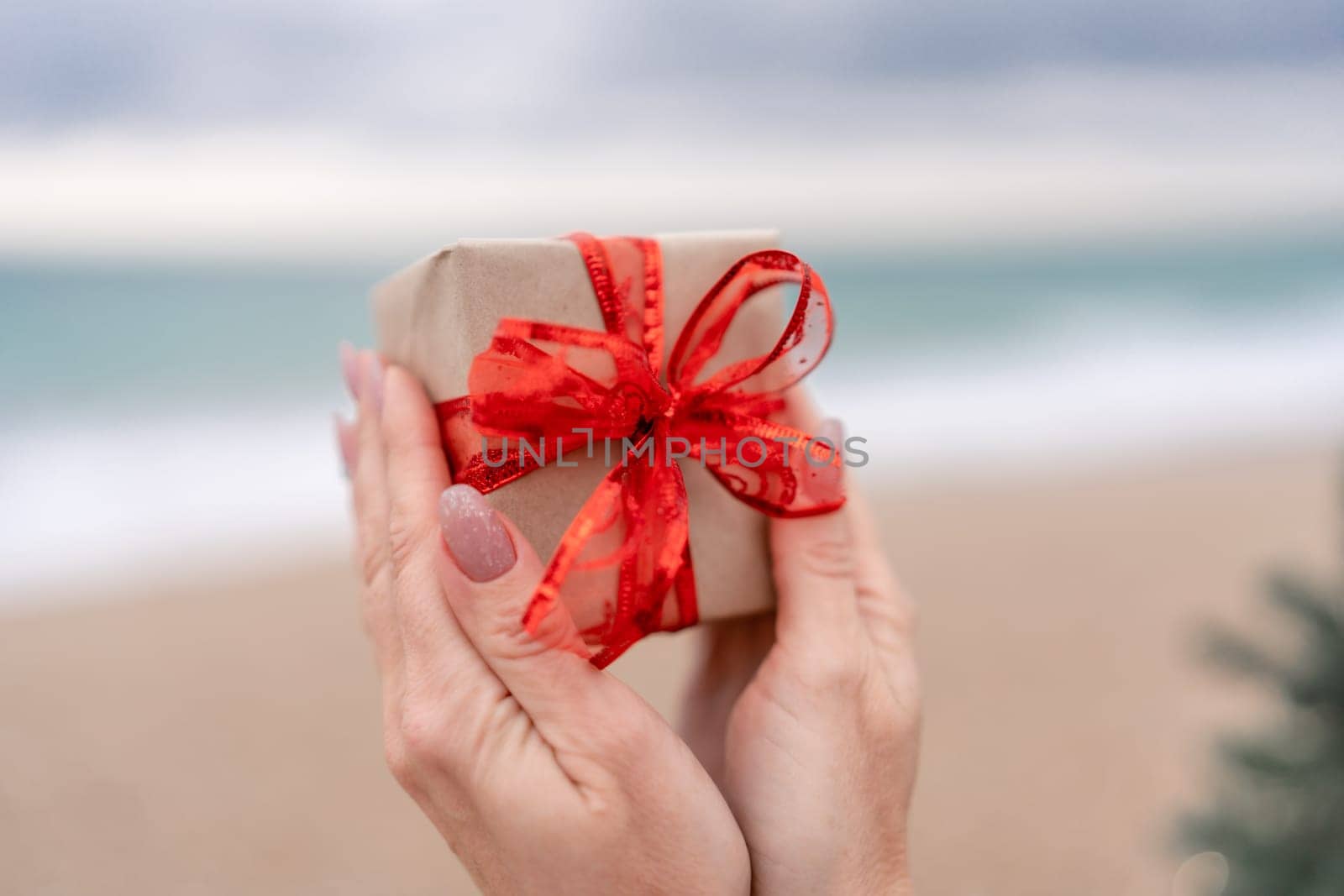 Hand holding wrapped gift with red bow. Sea Beach backdrop. The hand holding the gift is the main focus of the image, emphasizing the act of giving and receiving