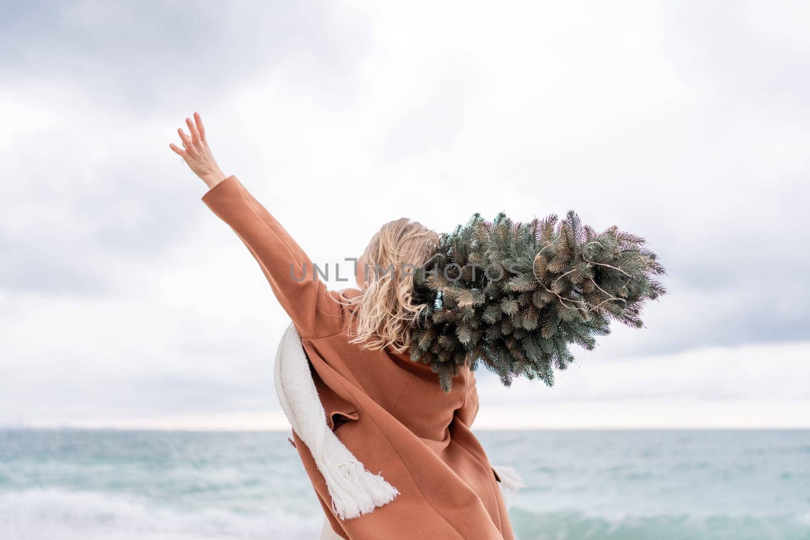 Blond woman Christmas sea. Christmas portrait of a happy woman walking along the beach and holding a Christmas tree on her shoulder. She is wearing a brown coat and a white suit