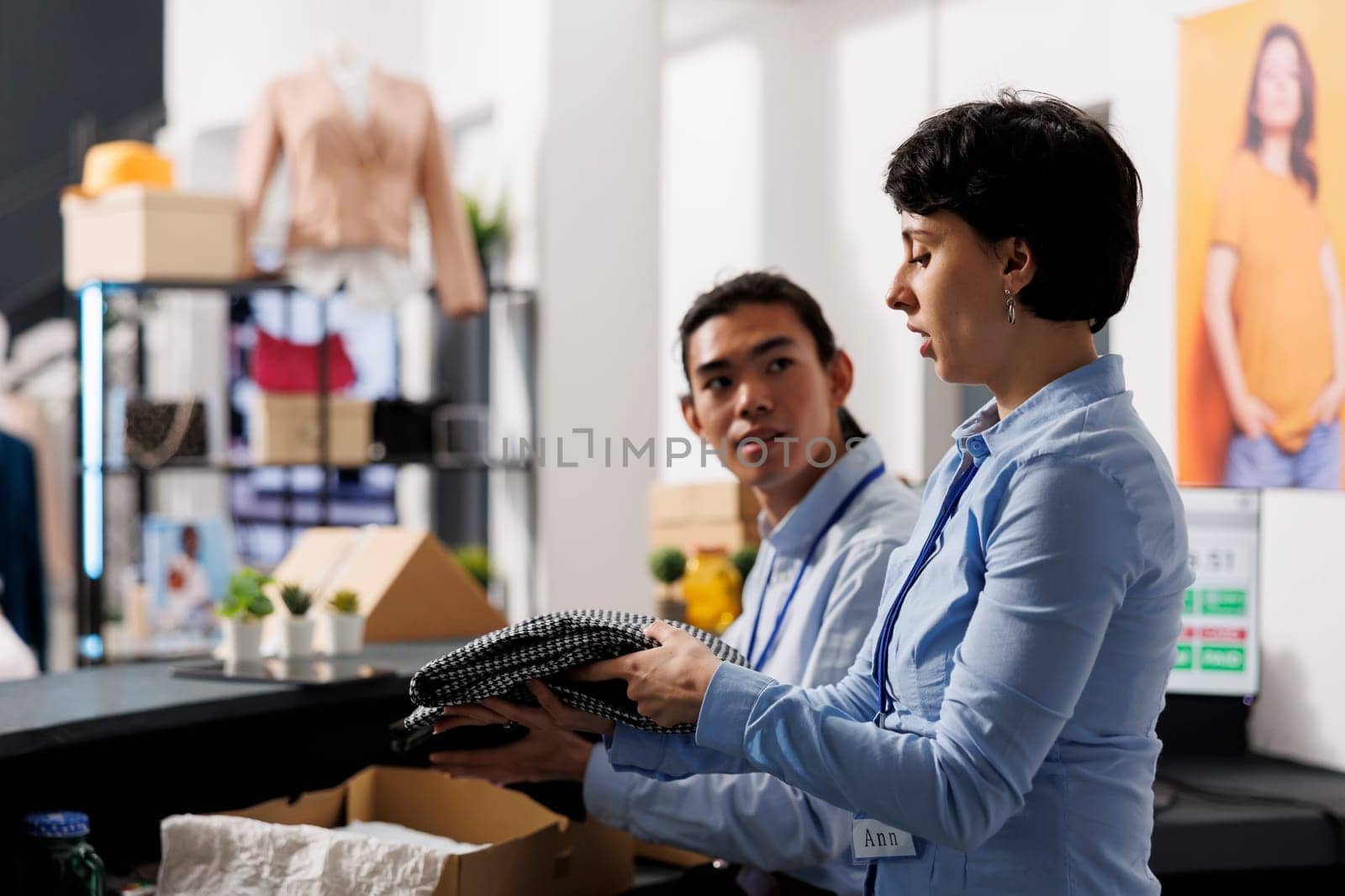 Store workers standing at counter desk, preparing packages for delivery in modern boutique. Woman putting stylish clothes in cardboard boxes, discussing shipping report with coworker in shopping mall
