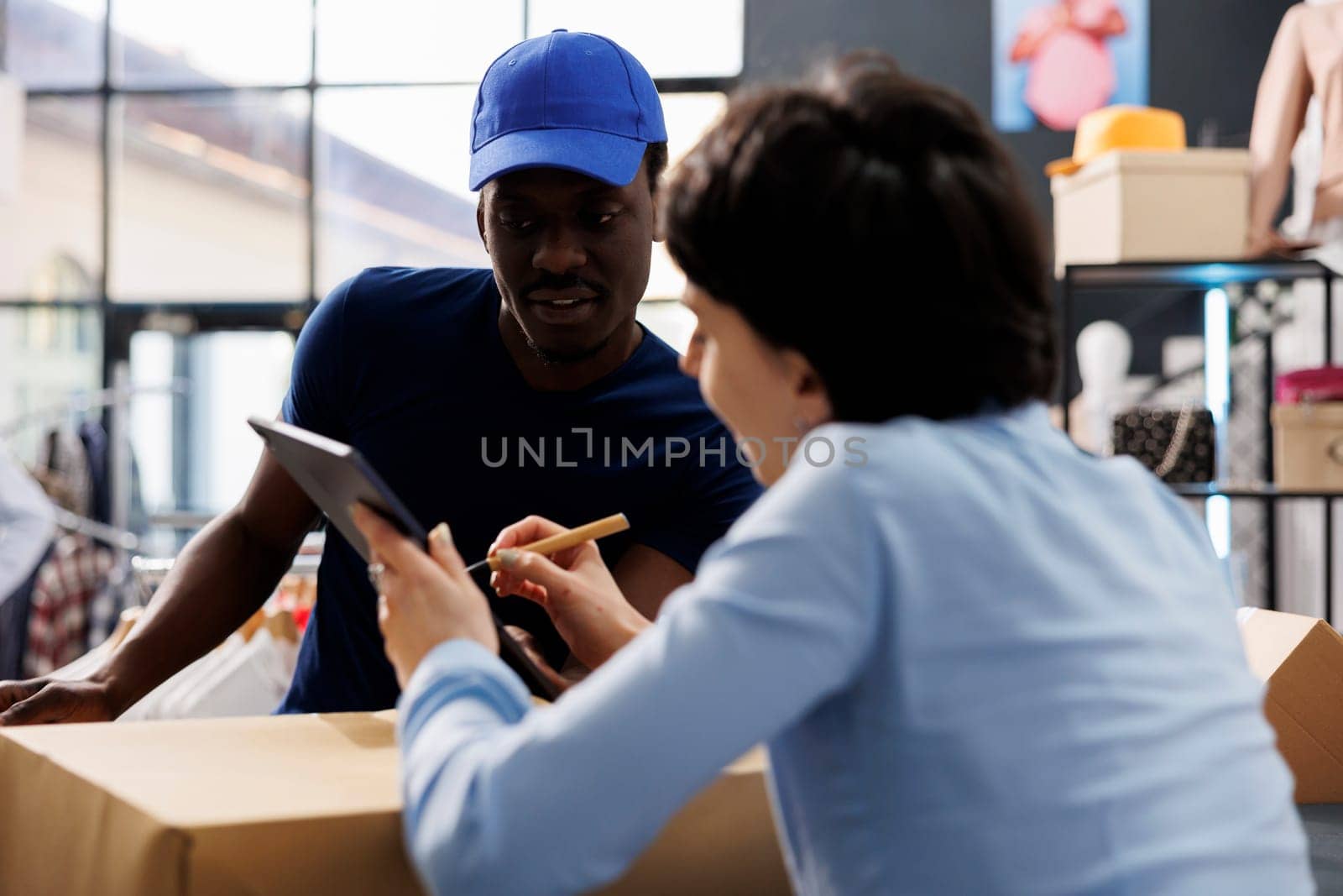 Store courier asking worker to sign delivery report, discussing customer shipping detalis in shopping centre. Stylish employee working at online orders, preparing packages in modern boutique
