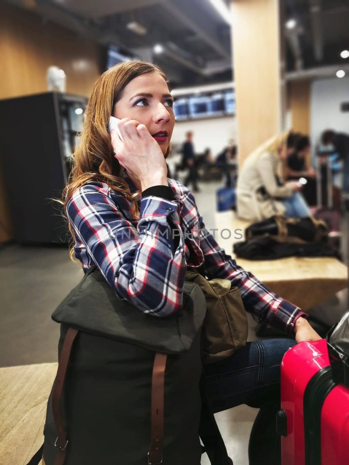 Young woman in shirt waiting for her transfer, with her backpack and luggage near her, talking on the phone, blurred station in background by Ivanko