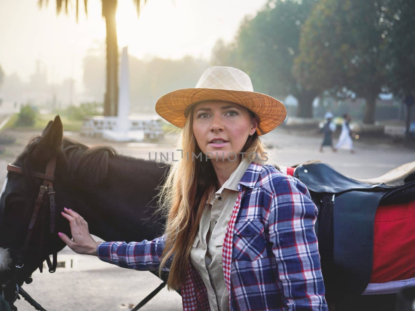 Candid portrait of young woman in stray hat and tartan shirt touching her black horse behind, ready for morning ride, blurred town in background