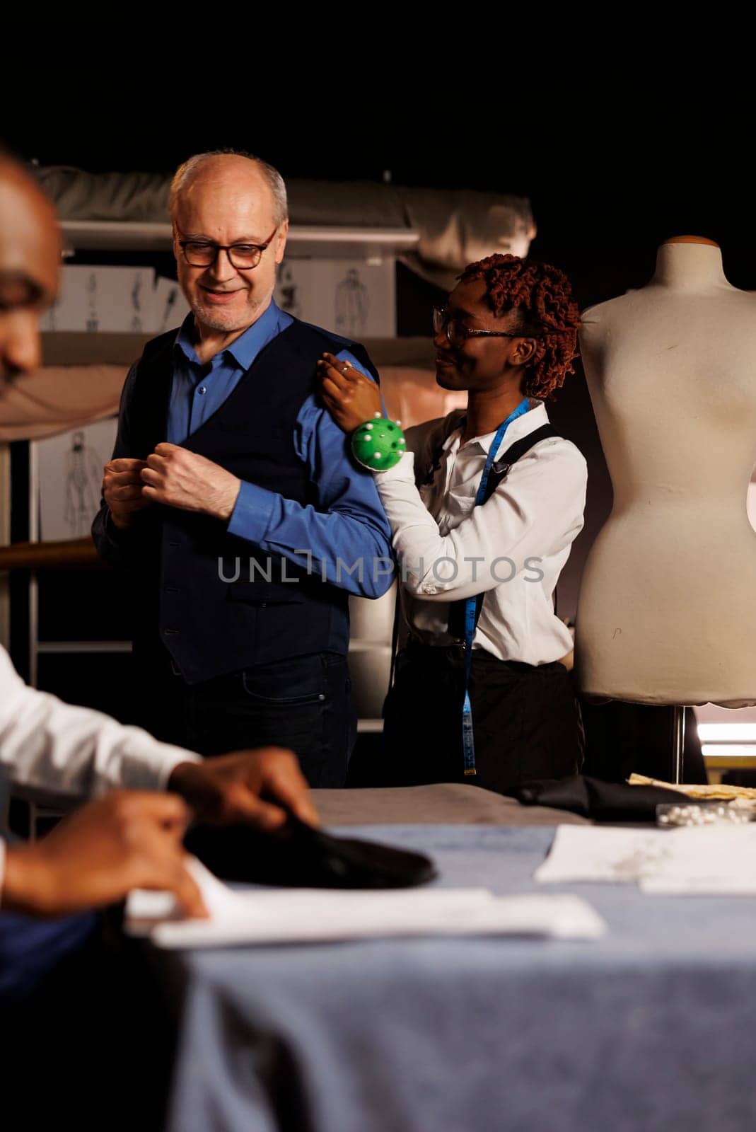 Cheerful meticulous seamstress doing precise additional needlework needed on sartorial piece worn by elderly client. Skilled african american tailor helping customer with bespoke suit comission