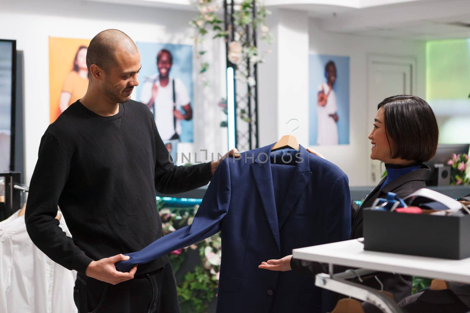 Smiling arab man holding jacket on hanger and getting fashion advice from clothing store asian woman assistant. Boutique client shopping for apparel while talking with consultant