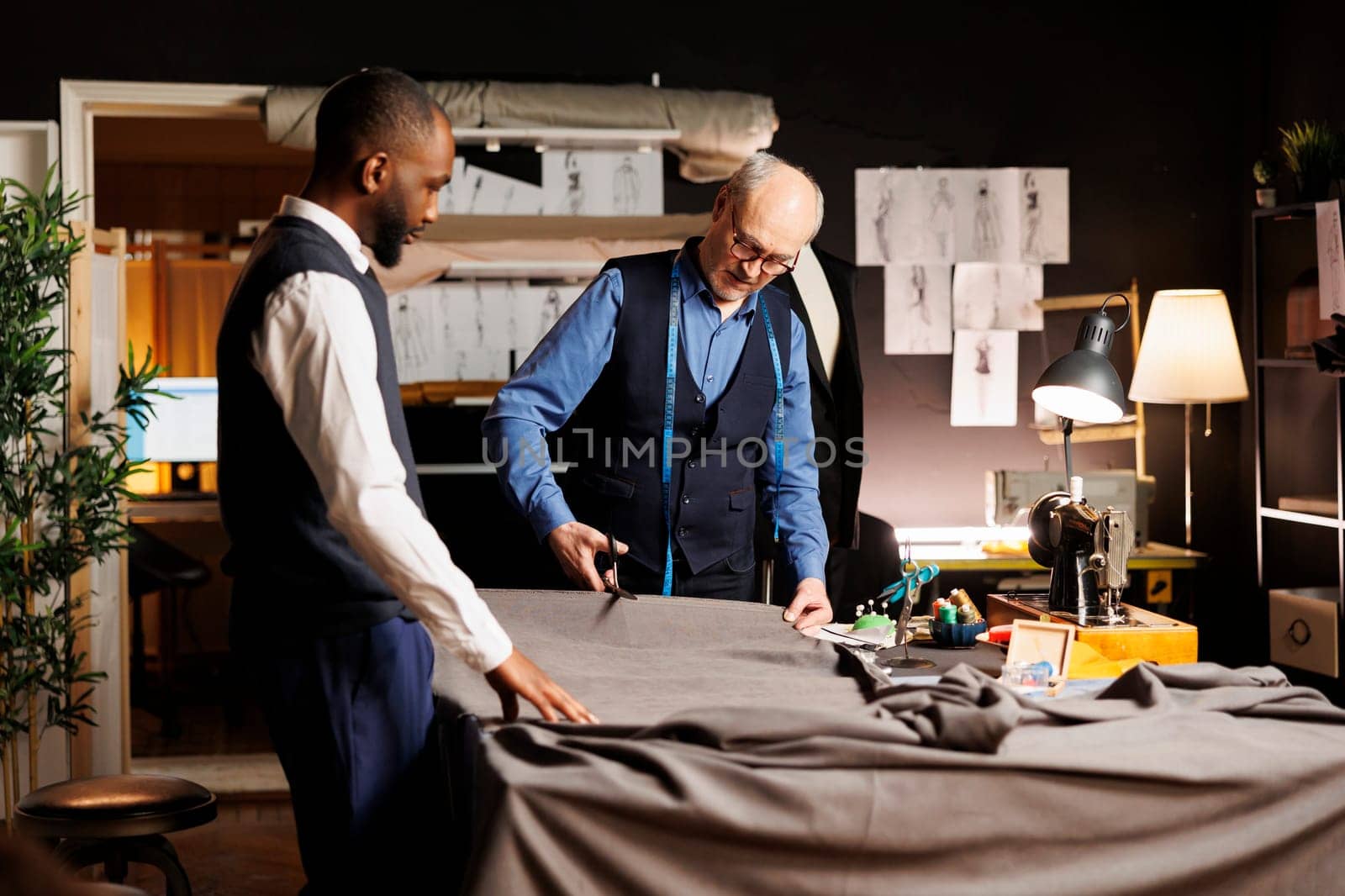 Meticulous elderly master tailor cutting fabric sheet for upcoming bespoke luxury fashion collection. Senior suitmaker showing african american apprentice expert craftmanship