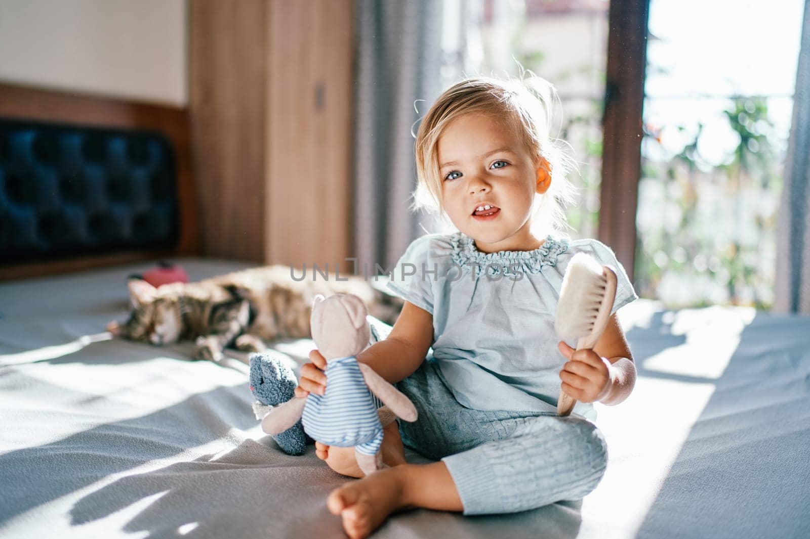 Little smiling girl sitting on the bed with a teddy bear and a comb in her hands. High quality photo